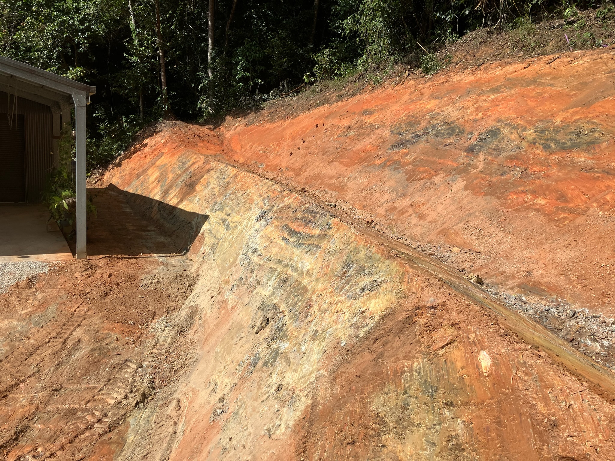 A steep batter and drainage swale cut beside a shed, rainforest above, shaped by Beau-Con Earthworks