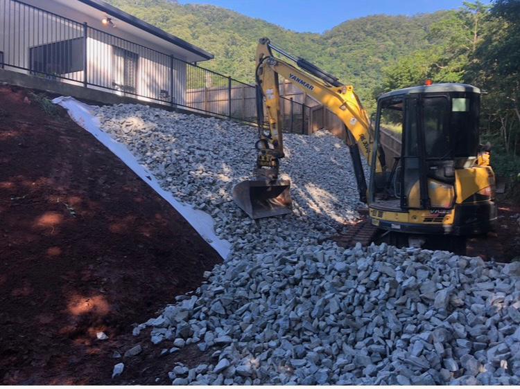 Rock protection placed over geotextile on a slope beside a home, a Yanmar excavator working the batter